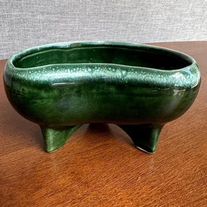 Puede incluir: Una jardinera de cerámica verde oscuro con acabado brillante. La jardinera de forma ovalada tiene tres pequeños pies triangulares. El borde tiene un aspecto verde más claro y moteado. Una pieza decorativa para plantas.