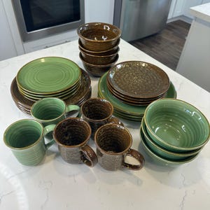 Op de afbeelding: Een set van groen en bruin keramiek serviesgoed, inclusief kommen, borden en mokken. De set heeft een gespikkelde bruine glazuur en een groene glazuur met een swirl-patroon.