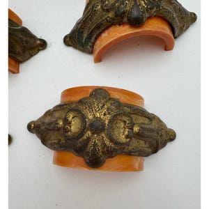 Puede incluir: Primer plano de varios tiradores de cajón antiguos de latón con un diseño ornamentado. El latón tiene un acabado dorado con detalles oscuros, montado sobre una base curva de color naranja. Los tiradores están dispuestos sobre una superficie blanca.