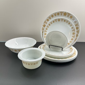 Puede incluir: Un conjunto de vajilla blanca y dorada con estampado floral. El juego incluye platos, cuencos y un plato pequeño. El diseño floral dorado decora los bordes de los platos y cuencos. La vajilla está dispuesta sobre una superficie oscura.