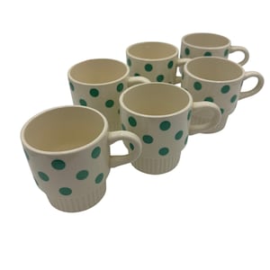 May include: Six cream-colored ceramic mugs with teal polka dots. Each mug has a handle and a stacked base. The mugs are arranged in a group, showcasing their design and shape. The interior of the mugs is a light cream color.