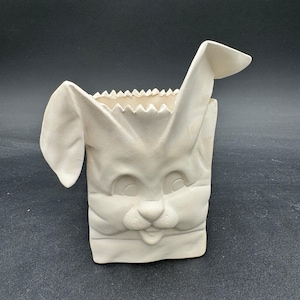 Könnte beinhalten: Ein keramischer, hasenförmiger Behälter in einer neutralen, cremefarbenen Farbe. Der Behälter ist so gestaltet, dass er einer Papiertüte mit Hasenohren und einem Hasengesicht ähnelt. Der obere Rand hat ein Zickzackmuster.