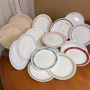 May include: A selection of white ceramic plates with patterned edges. Designs include hearts, floral motifs, and stripes in pink, blue, green, and red. Some plates are oval-shaped.