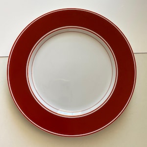 Puede incluir: Un plato redondo blanco con un borde rojo ancho. El borde rojo está acentuado con una fina línea blanca cerca del borde interior. El plato es limpio y sencillo, adecuado para el uso diario.
