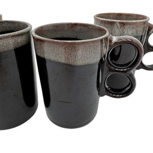 Puede incluir: Juego de cuatro tazas de cerámica negra con un diseño de asa único. Las tazas tienen una base marrón oscuro con un borde marrón más claro. Las asas presentan un diseño de estilo nudillo con tres orificios para los dedos.