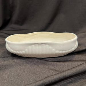 Puede incluir: Una jardinera ovalada de cerámica blanca con un diseño texturizado y acanalado alrededor del borde. El interior es de color crema claro. La jardinera está sobre un fondo oscuro.