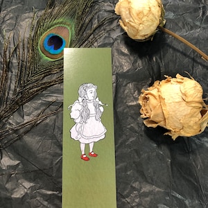 May include: A green bookmark with a black and white illustration of a girl with long hair wearing a white dress and red shoes.