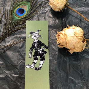 May include: A green bookmark featuring a black and white illustration of a scarecrow character. The bookmark is rectangular and set against a dark background with a peacock feather and dried roses.