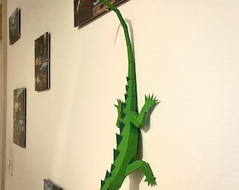 May include: A green papercraft iguana with red eyes is mounted on a white wall. The iguana's tail is curled up and its body is angled downwards.