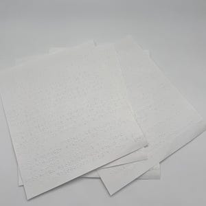 May include: A stack of white paper sheets featuring embossed Braille text. The tactile dots are arranged in horizontal lines across the surface of each rectangular page, creating a raised pattern designed for reading by touch.