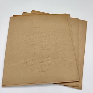 May include: A stack of rectangular, light brown paper sheets featuring embossed Braille text. The sheets are layered slightly offset, revealing multiple pages of tactile writing. The paper has a smooth, matte texture with visible indentations.