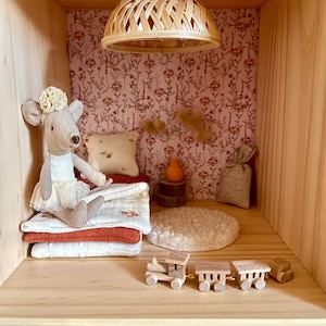 May include: A miniature dollhouse scene featuring a gray mouse doll wearing a dress, sitting on a stack of blankets. The scene includes a small wooden train, a pillow, and a woven lamp. The wallpaper has a floral pattern.