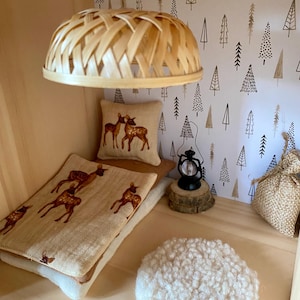 Puede incluir: Un dormitorio de casa de muñecas con una lámpara de tejido, una cama con un edredón y una almohada con estampado de cervatillo, una alfombra pequeña y una linterna sobre un tronco de árbol. La pared está cubierta con un papel tapiz blanco con un patrón de árboles marrones y negros.