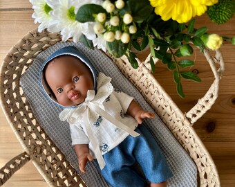 May include: A brown-skinned baby doll wearing a white and blue floral print shirt with a white lace collar and blue denim pants. The doll is lying in a woven wicker basket with a blue and white patterned fabric lining.