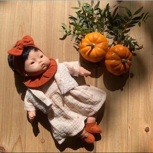 May include: A doll wearing a white dress with orange floral embroidery, a brown ruffled bib, and a brown headband with a bow. The doll is wearing brown shoes and is lying on a wooden surface with two orange pumpkins.