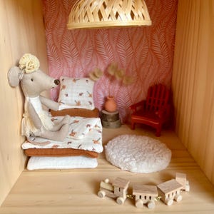 Può includere: Una scena di camera da letto di casa delle bambole in miniatura con una bambola di topo di peluche che riposa su un letto con biancheria da letto fantasia. La stanza presenta una sedia rossa, un piccolo treno di legno e un apparecchio di illuminazione intrecciato. Le pareti sono decorate con un motivo a foglie rosa e bianco.