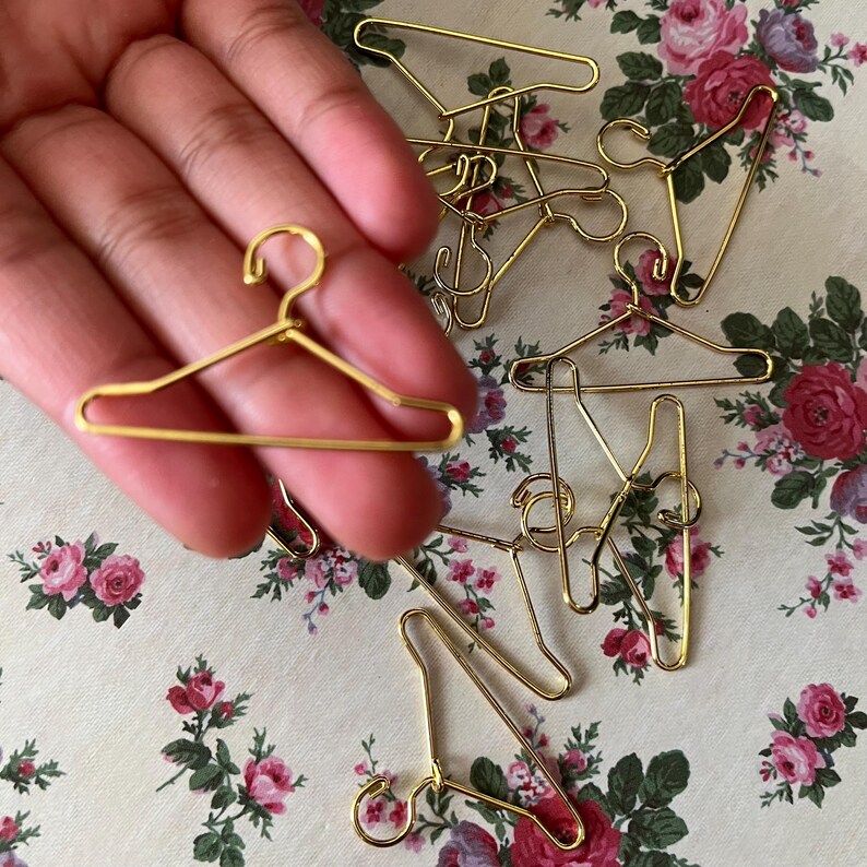 K&ouml;nnte beinhalten: Nahaufnahme einer Hand, die einen goldfarbenen Miniatur-Kleiderb&uuml;gel h&auml;lt. Der Kleiderb&uuml;gel ist aus d&uuml;nnem Draht gefertigt und hat ein schlichtes Design. Auf einer floral gemusterten Oberfl&auml;che liegen viele weitere Kleiderb&uuml;gel verstreut.
