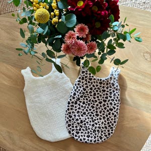 Puede incluir: Dos sacos de dormir para bebés. Uno es blanco con un patrón texturizado y punteado. El otro tiene un estampado de leopardo en blanco y negro. Los sacos de dormir están sobre una superficie de madera, con un ramo de flores en el fondo.