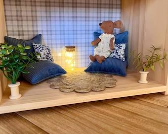 Könnte beinhalten: Ein Miniatur-Puppenhauszimmer mit einer karierten Tapete, zwei blauen Cordkissen, einem kleinen braunen Stofftier, das auf einem blauen Kissen sitzt, einer kleinen Topfpflanze und einer Lichterkette in einem Glas.