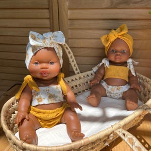 Può includere: Due bambole per bambini con tonalità della pelle scura sedute in un cestino di vimini. Una bambola indossa un completo giallo con fascia e top floreale. L'altra bambola indossa fascia e top floreale bianco con un completo giallo. Le bambole hanno tratti realistici.
