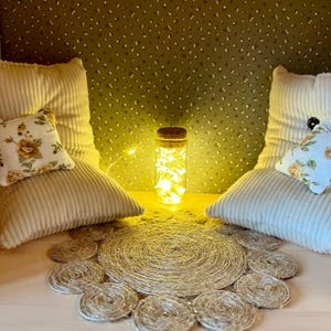 Puede incluir: Dos almohadas de pana blanca con almohadas de diseño floral sobre una superficie de madera clara. Un frasco de vidrio con luces de cadena se encuentra entre las almohadas sobre una alfombra redonda de yute tejido.