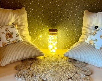Könnte beinhalten: Zwei weiße Cordkissen mit floralen Kissen auf einer hellen Holzoberfläche. Ein Glasgefäß mit Lichterketten steht zwischen den Kissen auf einem runden, gewebten Jute-Teppich.
