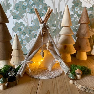 Può includere: Una tenda a forma di tipi in miniatura realizzata in tessuto beige con un cuscino bianco all'interno. Il tipi è decorato con perline di legno e una piuma. Il tipi è circondato da alberi di legno e un piccolo pezzo di muschio verde.