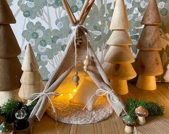 Könnte beinhalten: Ein Miniatur-Tipi-Zelt aus beigefarbenem Stoff mit einem weißen Kissen im Inneren. Das Tipi ist mit Holzperlen und einer Feder verziert. Das Tipi ist von Holzbäumen und einem kleinen Stück grünem Moos umgeben.