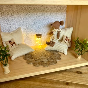 Puede incluir: Una habitación de casa de muñecas con una muñeca de ratón marrón sentada en una almohada blanca. La habitación tiene un papel tapiz con estampado blanco, una pequeña planta en maceta, una cadena de luces en un frasco de vidrio y una alfombra tejida.