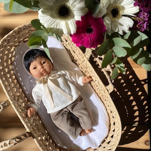 Peut inclure: Une poupée vêtue d'une chemise blanche, d'un pantalon marron et d'un bonnet, reposant dans un panier en osier. La poupée est allongée sur une couverture blanche à motifs. Le panier est entouré de fleurs blanches et roses et de feuilles vertes.