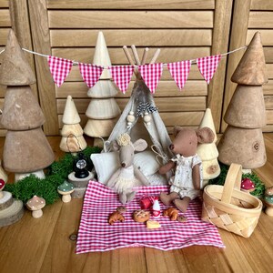 Peut inclure: Une scène de pique-nique fantaisiste avec deux poupées souris, une tente miniature et des arbres en bois. Une couverture à carreaux rouges et blancs contient de minuscules aliments. Une bannière à carreaux roses et blancs est suspendue au-dessus. Un panier en osier est à proximité.