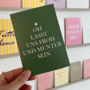Könnte beinhalten: Eine Hand hält eine dunkelgrüne Grußkarte mit weißem Text "OH LASST UNS FROH UND MUNTER SEIN" und einem Stern. Weitere Karten mit verschiedenen Farben und Texten sind im Hintergrund auf Regalen ausgestellt.