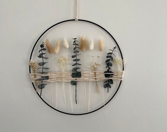 May include: A circular wall hanging featuring a black metal ring with dried flowers and greenery. The arrangement includes beige bunny tails, eucalyptus, and other dried floral elements, woven with natural twine. A neutral-toned decorative piece.