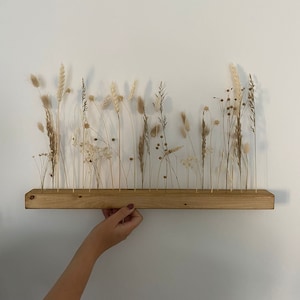 Op de afbeelding: Een houten plank met een rij gedroogde grassen en bloemen in verschillende tinten bruin en beige.