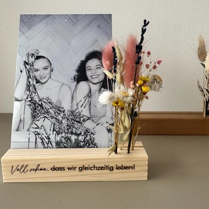 Op de afbeelding: Een houten fotohouder met een zwart-witfoto van twee vrouwen en een vaasje met droogbloemen. De houten basis heeft de tekst "Voll schön, dass wir gleichzeitig leben!". De bloemen zijn roze, geel en wit.