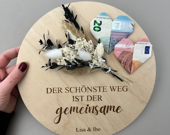 Peut inclure: Une pancarte ronde en bois avec le texte "Der schönste Weg ist der gemeinsame" et "Lisa & Ibo" écrit en brun. Deux cœurs en origami faits de billets d'euros sont attachés à la pancarte, ainsi qu'un bouquet de fleurs séchées.