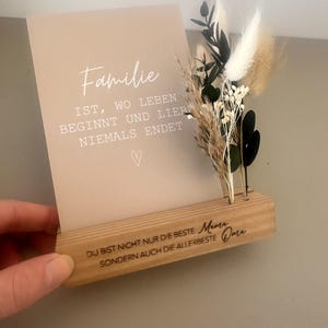 Könnte beinhalten: Ein Holz-Fotorahmen mit einer beigefarbenen Karte mit der Aufschrift "Familie" und einem Zitat auf Deutsch. Der Halter hat ein kleines Trockenblumenarrangement. Die Basis des Halters hat gravierten Text.