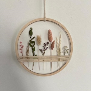 Op de afbeelding: Een houten hoepel met gedroogde bloemen en groen geordend erin. De hoepel hangt aan een touwtje en heeft een geweven band van touw in het midden.