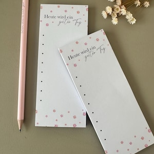 Könnte beinhalten: Ein rosa Bleistift und zwei weiße Notizblöcke mit dem Text "Heute wird ein guter Tag" in schwarzer Schrift. Die Notizblöcke haben ein Blumendesign mit kleinen rosa Blüten und schwarzen Punkten. Der Bleistift hat Text in einer Schriftart.