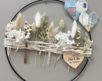 May include: A circular wall hanging with dried flowers and origami hearts made from currency. The black metal ring is adorned with white and beige dried flowers, wrapped with twine. Two origami hearts are visible, and a wooden heart tag reads "Lisa & Ibo 26.10.2024".