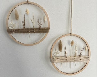Puede incluir: Dos colgantes de pared circulares de madera con flores y hierbas secas. Los aros de madera natural están envueltos con hilo y suspendidos con hilo. Los arreglos florales secos incluyen elementos beige, marrones y verdes.