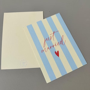 Könnte beinhalten: Ein cremefarbenes Karten- und Umschlag-Set. Die Karte hat blaue und weiße vertikale Streifen und die Worte "just married" in roter Schrift mit einem kleinen roten Herz. Der Umschlag ist schlicht.