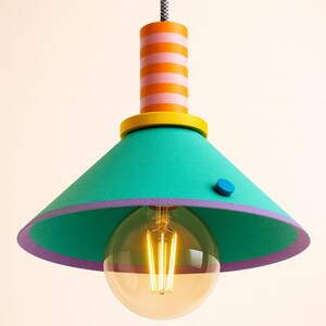 Op de afbeelding: Een hanglamp met een conische turquoise kap, een paarse rand en een klein blauw rond accent. Het cilindrische bovendeel heeft horizontale oranje en roze strepen, bekroond door een gele ring, met een grote ronde gloeilamp eronder.