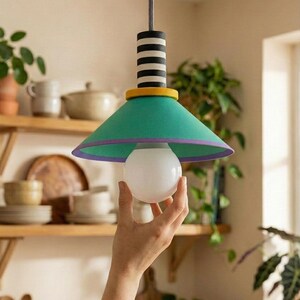 Op de afbeelding: Een hanglamp met een conische groene kap met een paarse rand, een geel cirkelvormig accent en een zwart-wit gestreept cilindrisch bovendeel. De lamp hangt aan een getextureerd snoer en is voorzien van een ronde witte gloeilamp.