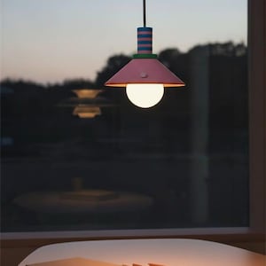 Op de afbeelding: Een hanglamp met een conische roze kap, een groene accentring en een cilindervormig bovendeel met horizontale blauwe en roze strepen. Een ronde, wit oplichtende lamp hangt aan een snoer vanuit het midden van de kap naar beneden.
