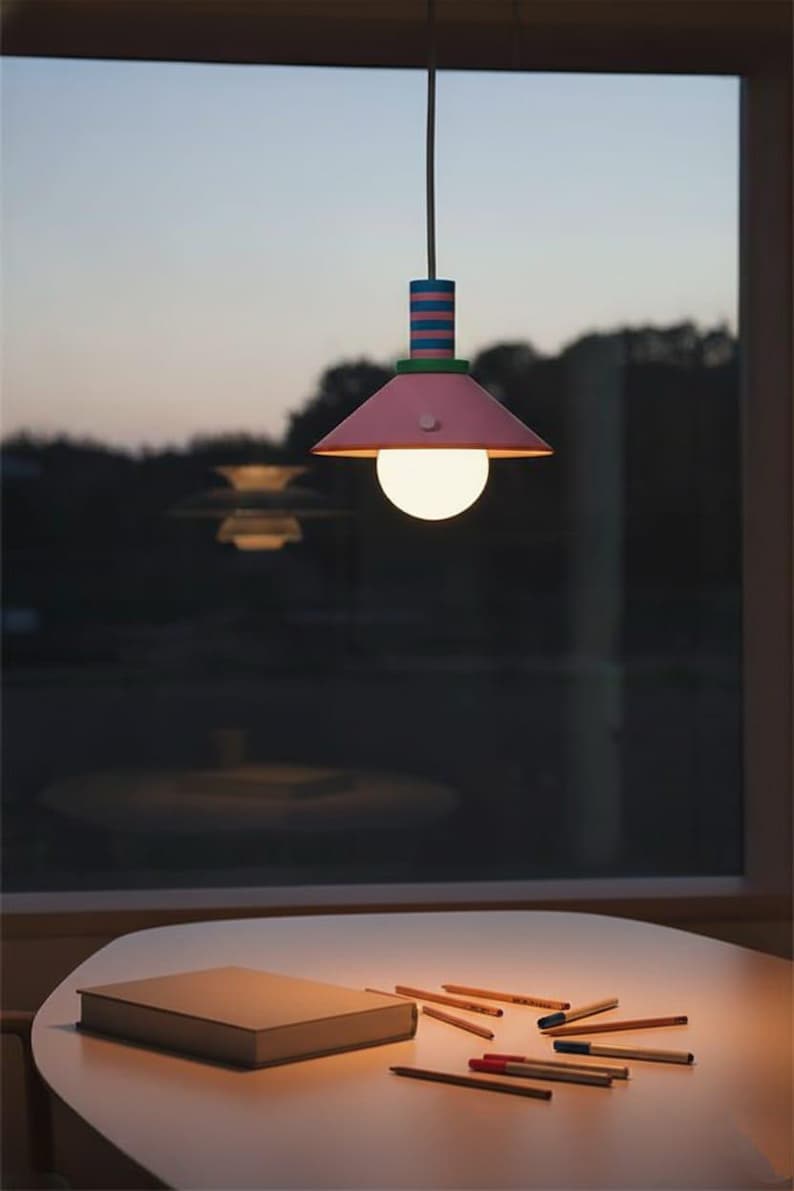 Op de afbeelding: Een hanglamp met een conische roze kap, een groene accentring en een cilindervormig bovendeel met horizontale blauwe en roze strepen. Een ronde, wit oplichtende lamp hangt aan een snoer vanuit het midden van de kap naar beneden.
