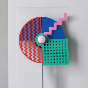 Könnte beinhalten: Eine farbenfrohe, geometrische Wanduhr im Retro-Design. Die Uhr hat einen roten und blauen Halbkreis, einen blauen Kreis, ein grünes Quadrat mit Gittermuster und eine rosa Treppe. In der Mitte befindet sich eine weiße Kugel. Ein silberner Draht hängt darunter.