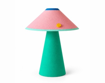 Op de afbeelding: Een tafellamp met een conische groene voet en een brede, conische roze lampenkap. De kap is voorzien van een blauwe bovenrand, een groenblauwe onderrand en een kleine, ronde gele accentknop op de roze zijde.