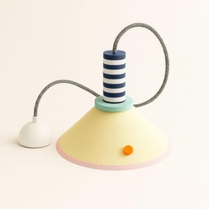 Op de afbeelding: Een hanglamp met een conische lichtgele kap met een roze onderrand en een klein oranje cirkelvormig accent. Het heeft een cilindrisch bovendeel met marineblauwe en witte horizontale strepen, een mintgroene basis en een zwart-wit snoer.