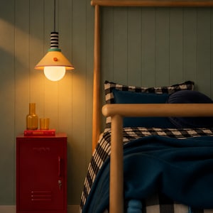 Op de afbeelding: Een hanglamp met een conische gele kap, een zwart-wit gestreepte hals en een bolvormige witte lamp. De armatuur hangt boven een rood metalen nachtkastje, waarop een glazen fles, een glazen beker en een rood boek zijn geplaatst.